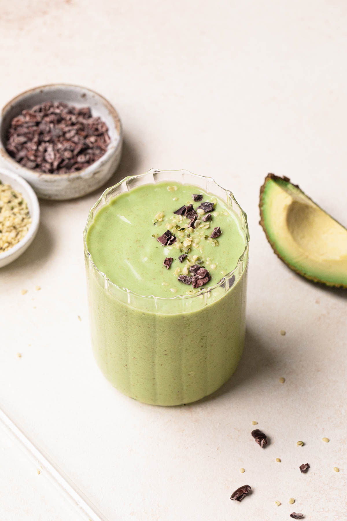 Green smoothie in a clear glass topped with hemp seeds and cacao nibs, avocado wedge