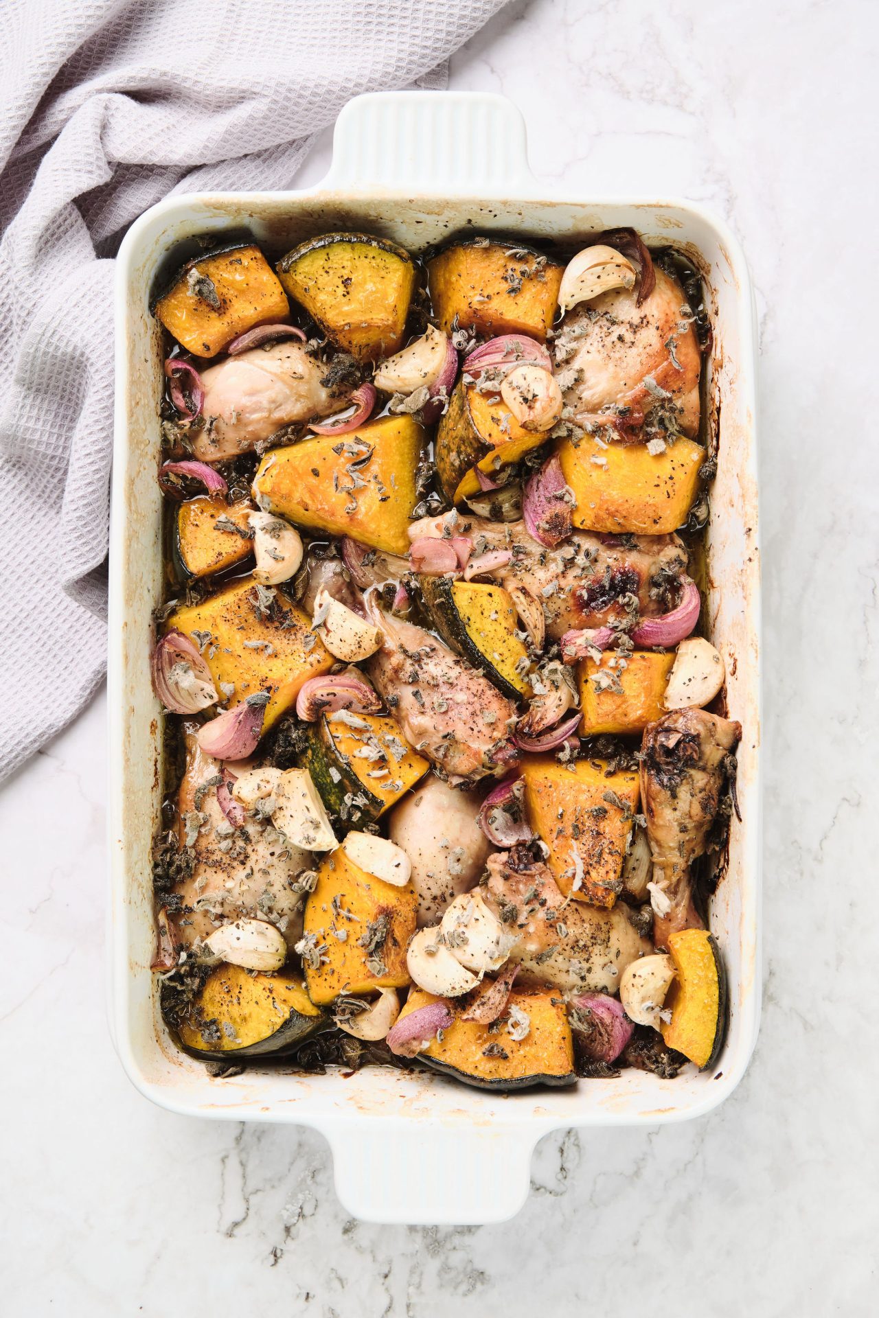 Roasted chicken in a white baking dish with pumpkin, onion and herbs