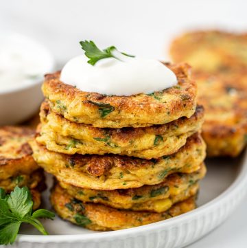 Stack of gluten free corn fritters on a white plate topped with yoghurt and herbs.