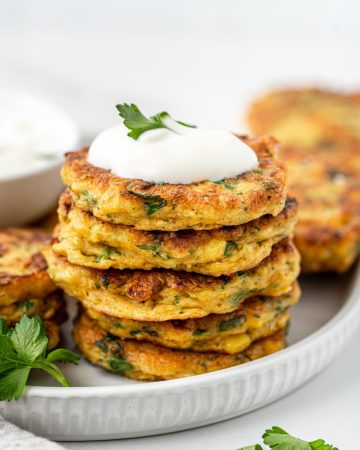 Stack of gluten free corn fritters on a white plate topped with yoghurt and herbs.