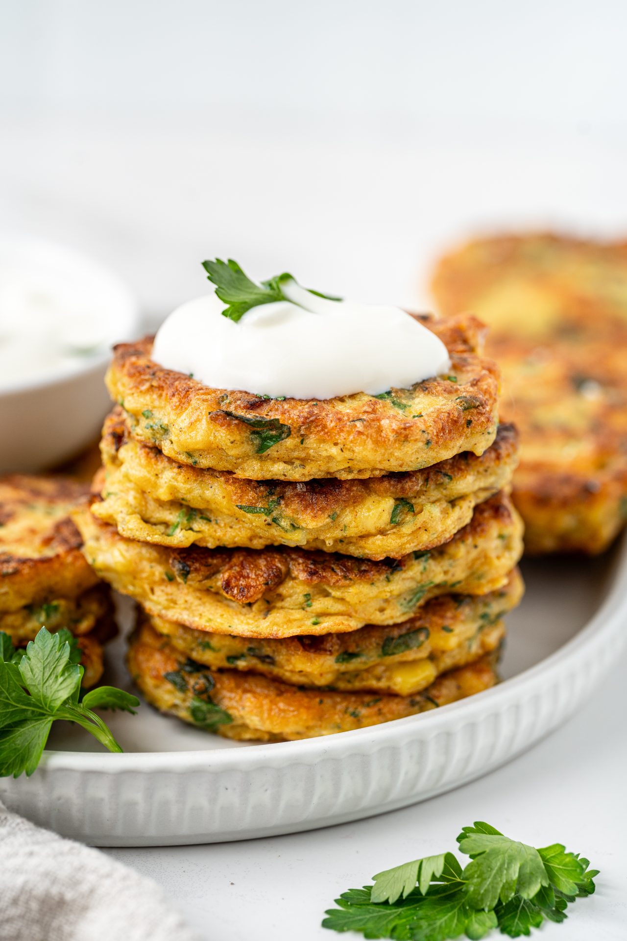 Stack of gluten free corn fritters on a white plate topped with yoghurt and herbs.