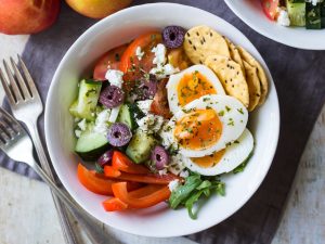 Meal prep breakfast bowls Greek style, combining boiled eggs with chopped cucumber, tomatoes, capsicum, feta and olives.