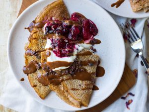 Sweet vegan buckwheat crepes folded on white plate served with yoghurt, berries and chocolate sauce.